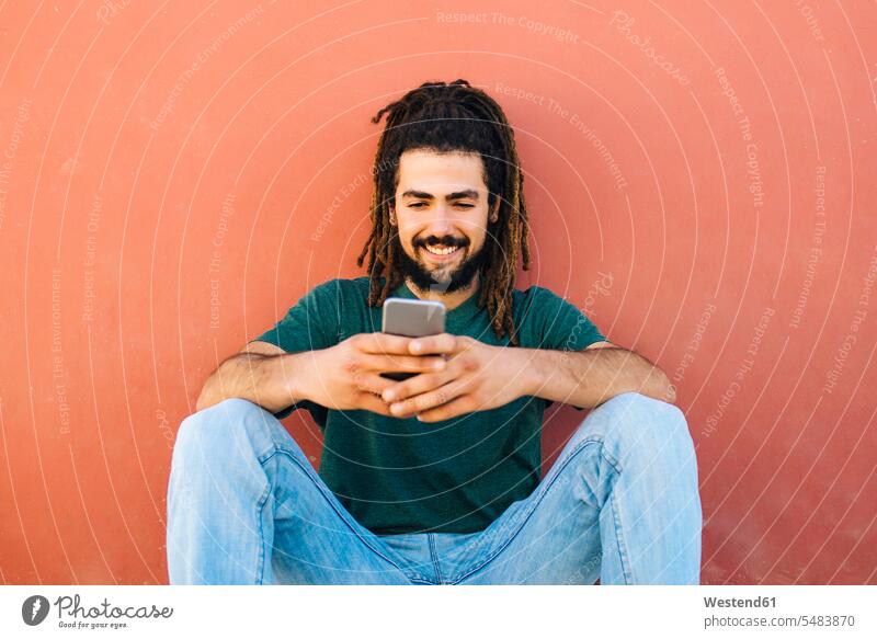 Portrait of smiling young man with dreadlocks and beard looking at his smartphone in front of a reddish wall style stylish front view frontal View From Front