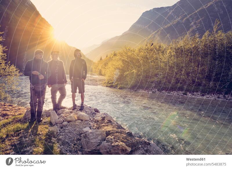 Slovenia, Bovec, three friends at Soca river at sunset River Rivers man men males standing water waters body of water Adults grown-ups grownups adult people