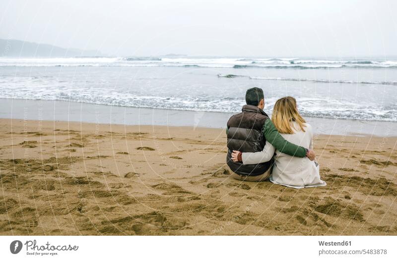 Couple in love sitting on the beach in winter beaches Seated couple twosomes partnership couples people persons human being humans human beings ocean water