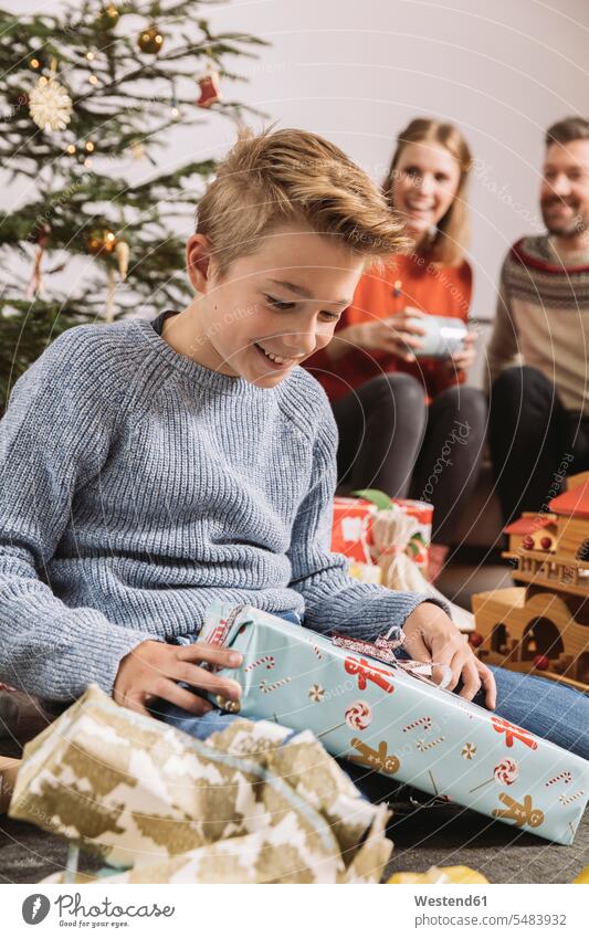 Little boy unwrapping a Christmas gift, parents sitting on couch in background caucasian caucasian ethnicity caucasian appearance european Joy enjoyment