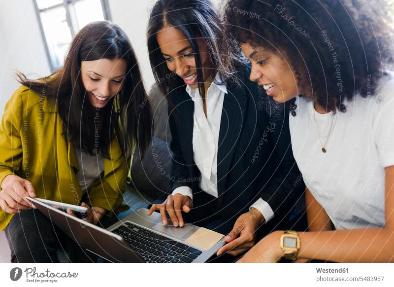 Three smiling businesswomen sharing laptop in office offices office room office rooms workplace work place place of work businesswoman business woman