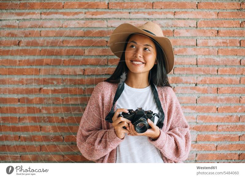 Portrait of smiling young woman with camera in front of brick wall cameras females women Adults grown-ups grownups adult people persons human being humans