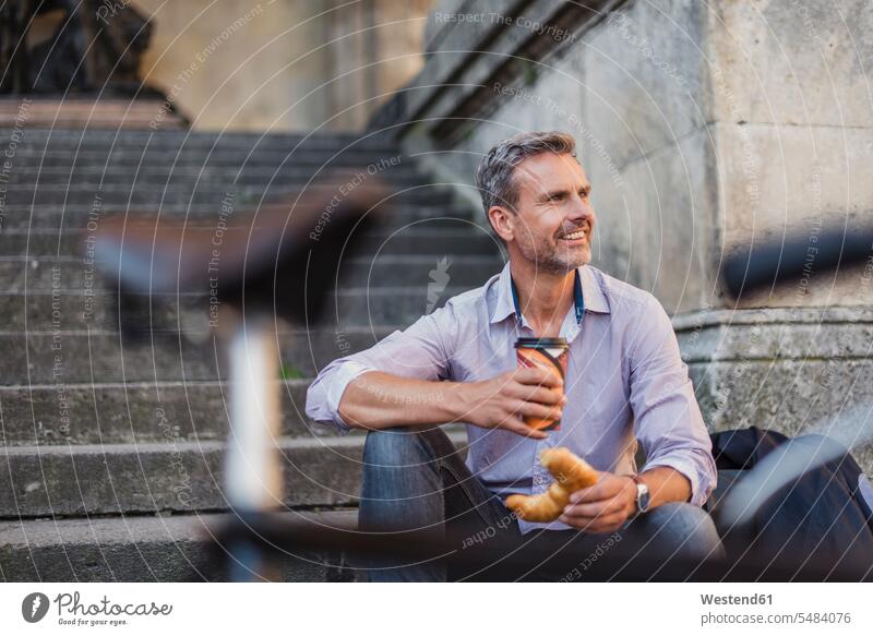 Smiling man sitting on stairs with croissant and takeaway coffee in the city smiling smile men males bicycle bikes bicycles Seated Coffee town cities towns