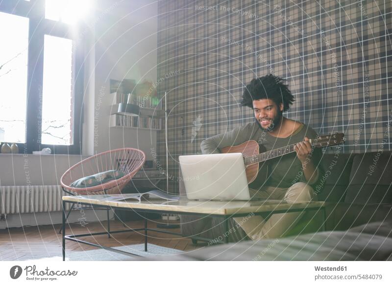 Man sitting in living room on sofa playing guitar in front of laptop guitars man men males Seated Laptop Computers laptops notebook stringed instrument
