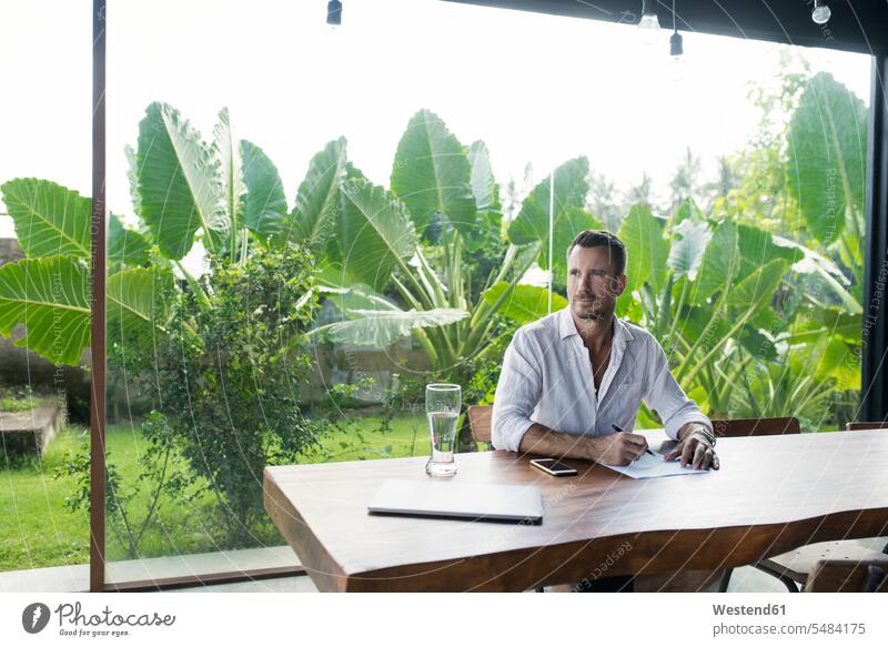 Mature man sitting at table in front of lush garden, writing gardens domestic garden mature men mature man thinking Seated write working At Work Table Tables