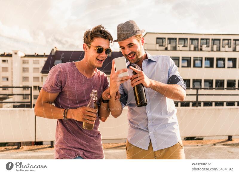 Two friends with beer bottles and cell phone on rooftop roof terrace deck mobile phone mobiles mobile phones Cellphone cell phones Fun having fun funny laughing