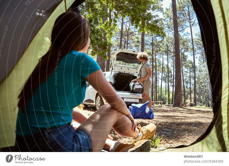 Young woman sitting in a tent watching her boyfriend packing the car tents loading automobile Auto cars motorcars Automobiles couple twosomes partnership