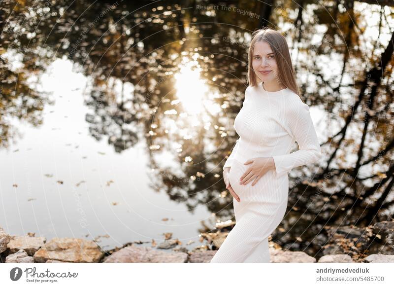 Mothers day. Beautiful young pregnant woman in white tight knit dress with little belly relaxing outdoors by the pond with reflection nature and sun in the park and looking at camera. Women day