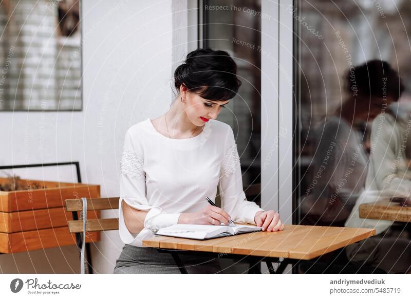 Serious focused smart businesswoman is writing notes on notepad, making agenda on personal organizer. Female in business suit sitting at table at cafe on terrace, work and event planning.