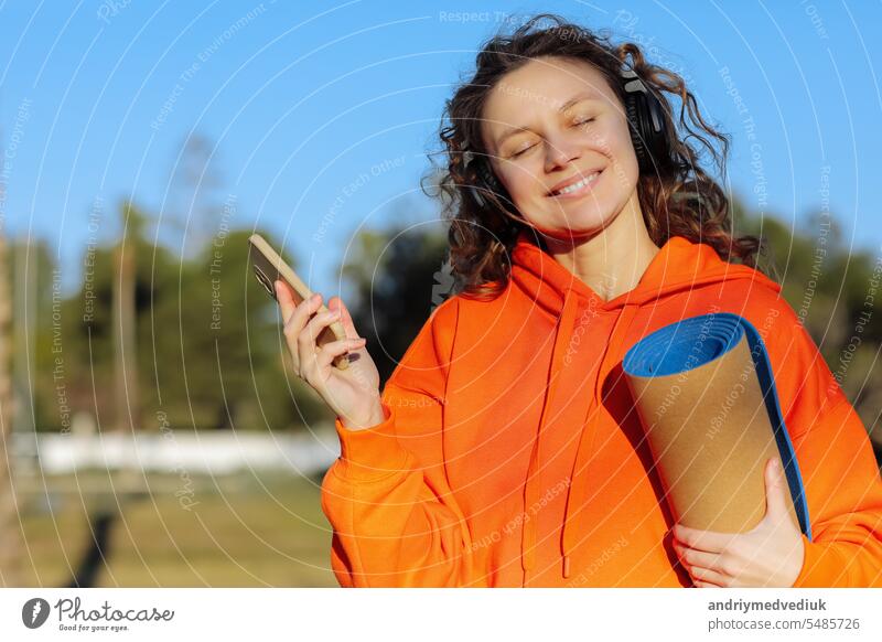 Happy young woman In headphones with yoga mat, listens music on smartphone outdoors. Smiling girl with closed eyes in vivid orange sweatshirt enjoys favorite playlist after training in green park