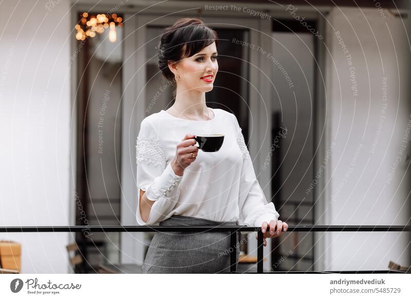 Young beautiful woman holding a cup of coffee or tea in cafe, restaurant. Brunette girl is drinking hot drinks, looking in camera outdoors on terrace and having lunch break during work. copy space