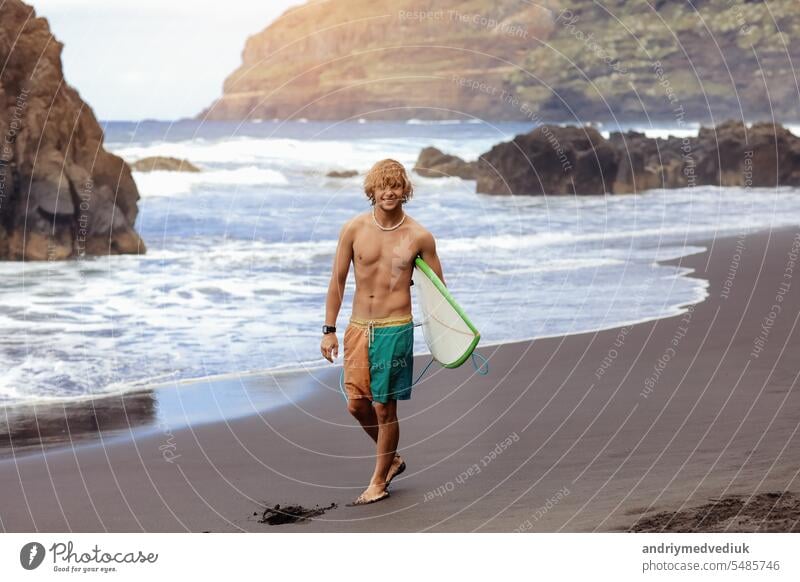 Fit young surfer man with curly blond hair with surfboard goes by the ocean having fun doing extreme water sports, surfing. Travel and healthy lifestyle concept. Sports travel destination.