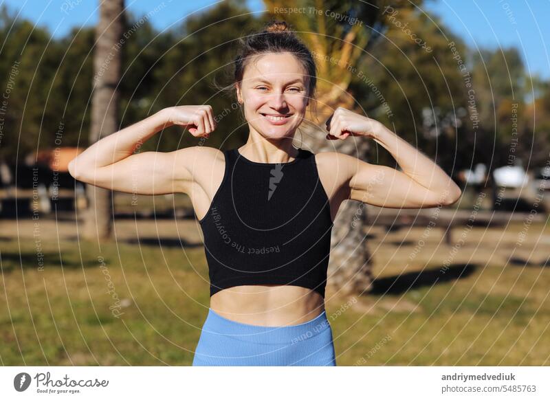 Young beautiful woman in sportswear raises arms to show muscles, biceps, demonstrates strong hand, has slim figure outdoors, feels confident in victory, looks independent, smiles positively