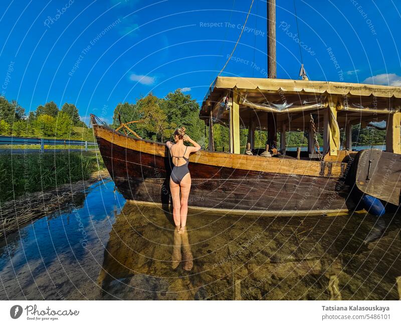 A woman is resting in the lake standing near a vintage wooden ship. Attractive fit woman is resting near the lake in summer. slim swimsuit water old college