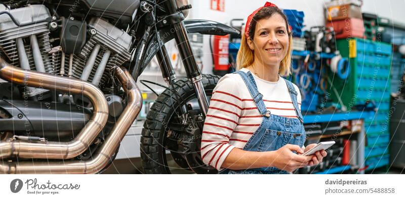 Mechanic woman holding phone sitting over platform with motorcycle on factory mechanic portrait female smiling happiness motorbike using cellphone looking