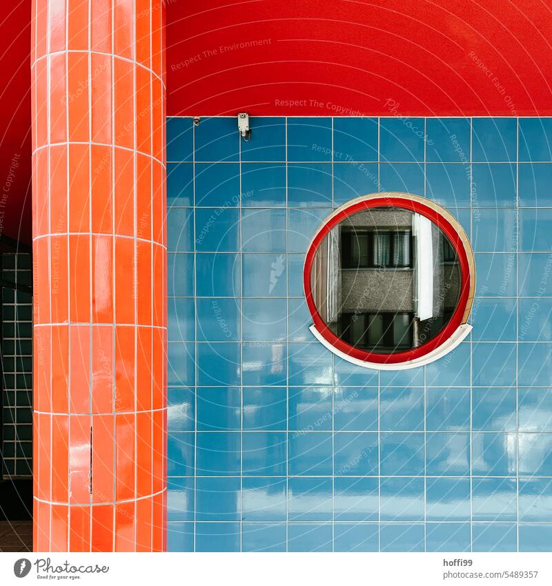 Colorful into the day - facade with blue, red, orange tiles and round window reflecting a gray facade, as well as photographer and a few cars. Blue Red Orange