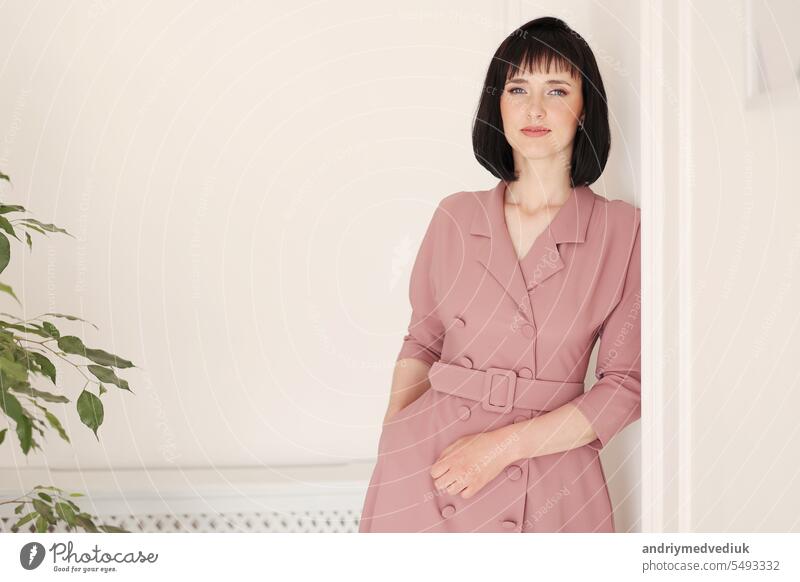 Portrait of attractive smiling brunette with short hair woman staying alone on white stylish wall background with green houseplant. Female in soft pink dress is looking in camera