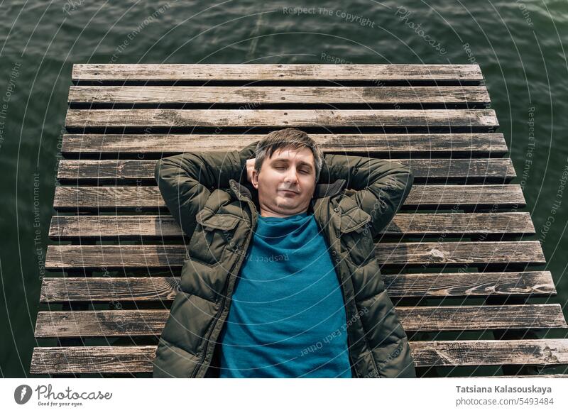 A man lies on a wooden plank pier on the lake in a jacket and jumper with his eyes closed Man relaxing boat dock enjoying sunny autumn day relaxes lying resting