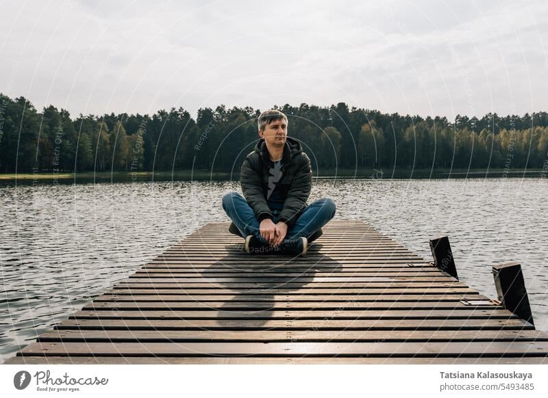 A man enjoys the view while exploring nature while sitting cross-legged on a wooden pier in autumn jeans jumper jacket relaxing boat dock lake enjoying sunny