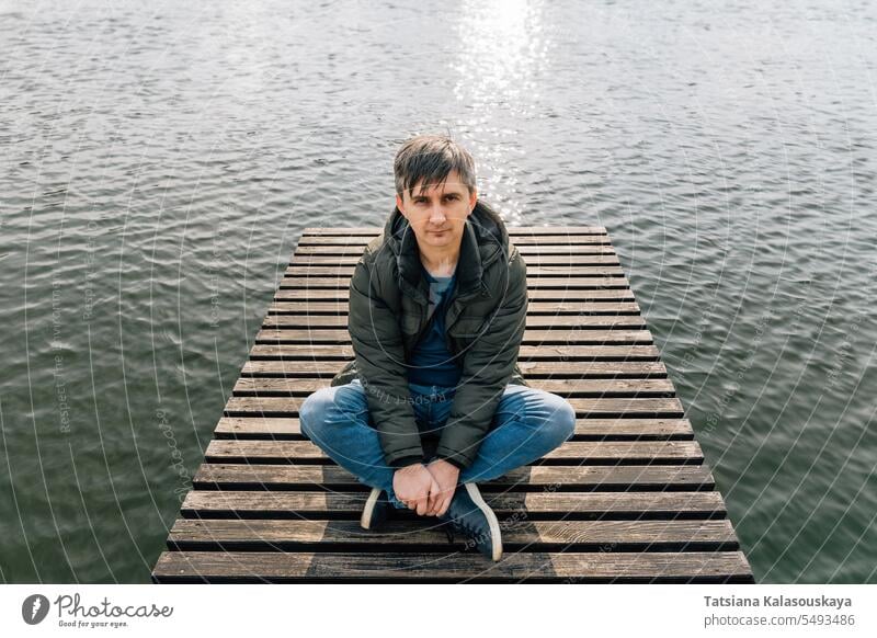 A man sits relaxed on a wooden pier on the lake, cross-legged in jeans and a jacket in autumn jumper relaxing boat dock enjoying sunny day relaxes sitting
