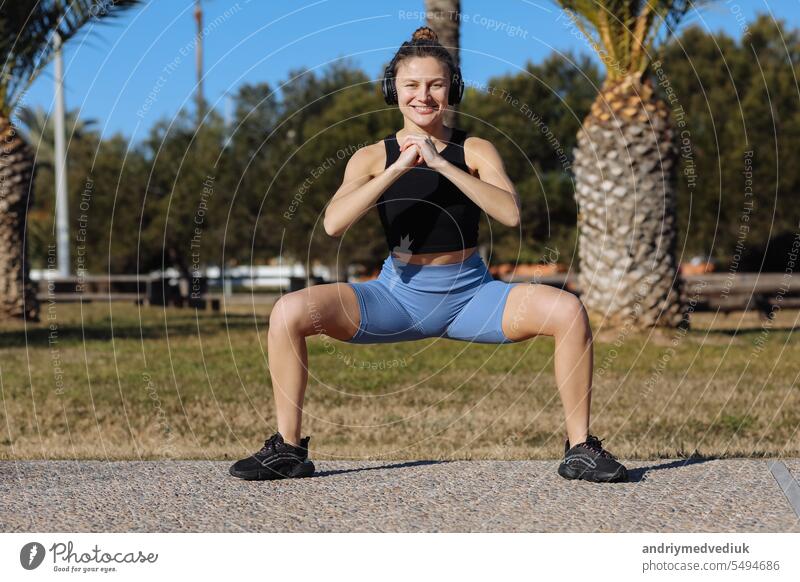 Young healthy woman in sportswear listens music in headphones while doing fitness squat leg exercises outdoors in park with palm trees. Workout for strengthen pelvic floor muscles on summer holiday