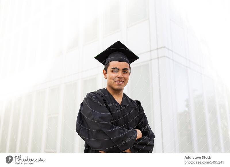 Man in graduation gown and cap standing against wall man young arms crossed confident student achievement portrait education handsome black white celebration