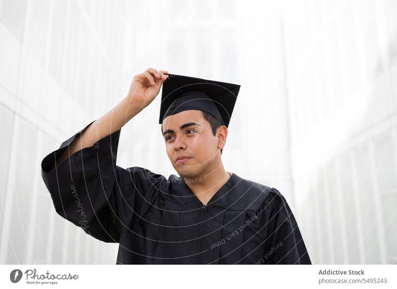 Man in graduation gown and cap on a white background looking away man young confident student achievement portrait education handsome black wall standing