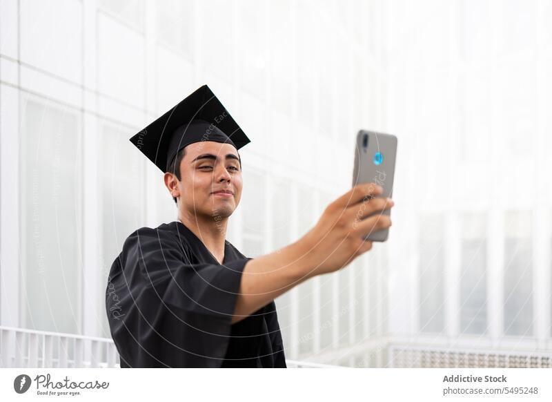 Man in graduation gown and cap taking selfie over phone man mortarboard smartphone student gadget achievement smile handsome mobile black cellphone degree