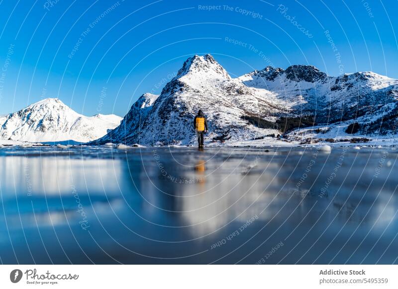 Low angle traveler standing on frozen lake near snowy mountains tourist winter nature lofoten island norway shore coast cold blue sky ice scenic north trip