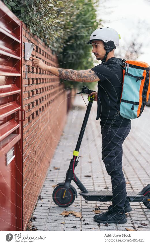 Courier man ringing doorbell of house in neighborhood delivery man electric kick scooter transport service gate courier modern helmet urban vehicle daytime