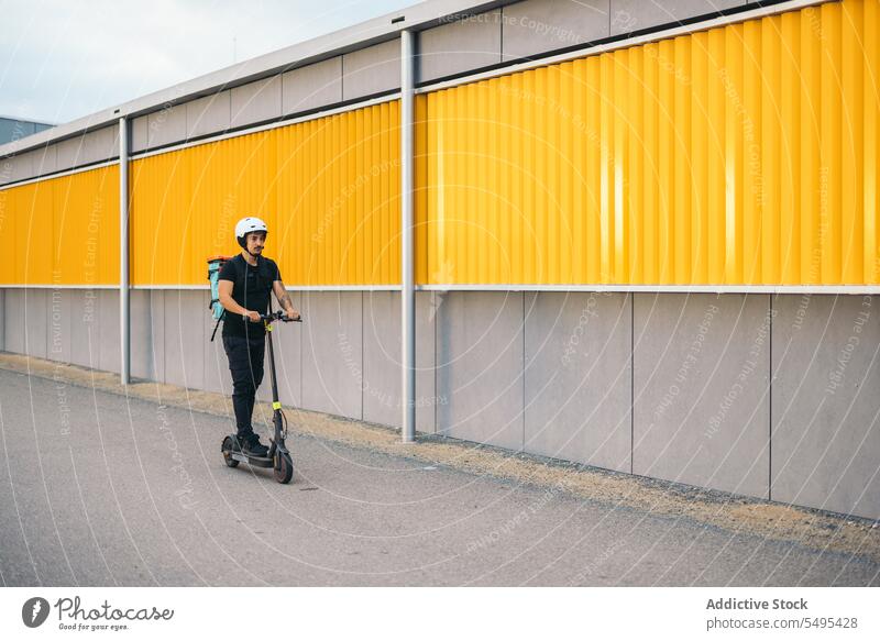 Courier service worker riding electric scooter on road delivery man kick scooter ride transport courier city street modern building vehicle male wall asphalt