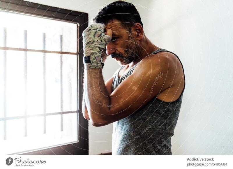 Strong adult man boxer with hands wrapped in cloth standing near window in room muscular workout athlete sport thoughtful strong exercise male confident fit