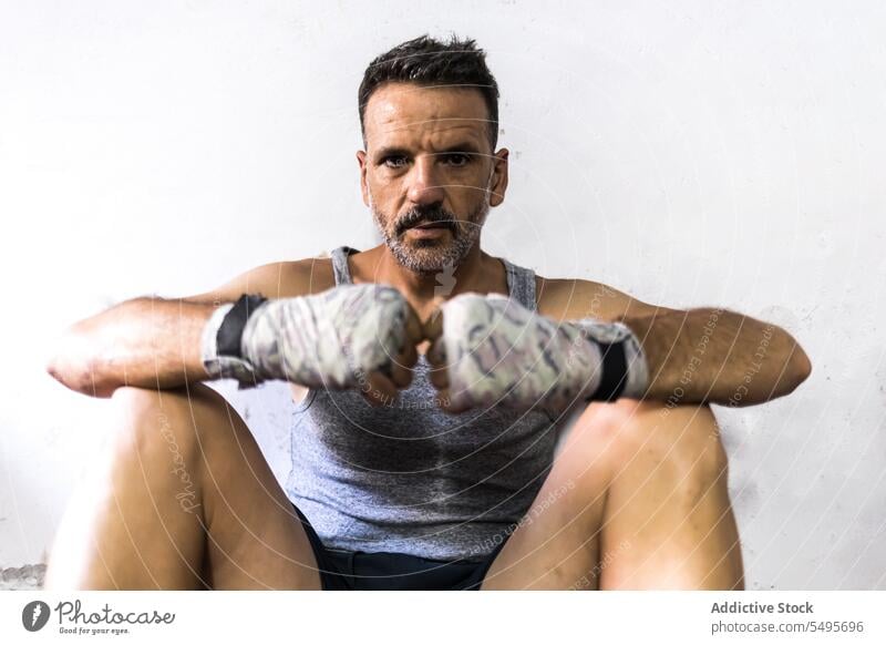 Strong adult man sitting on floor with wrapped hands in daylight thoughtful calm serious room confident athlete wall lean on male pensive individuality
