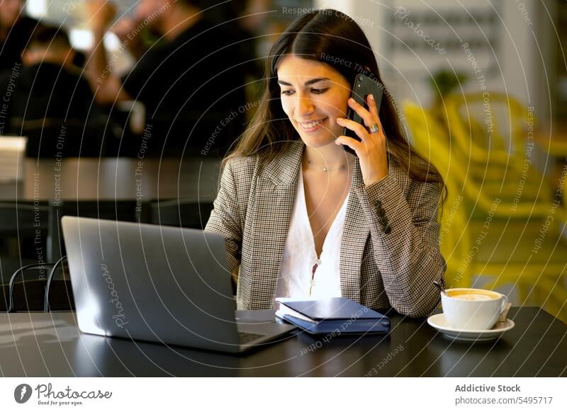 Smiling woman talking on smartphone while browsing laptop in cafe phone call freelance remote work device speak conversation using netbook mobile coffee gadget
