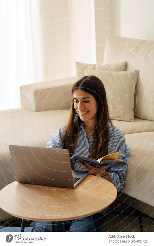 Happy young woman reading book while sitting with laptop near sofa positive free time hobby smile comfort at home female device gadget happy cheerful lady