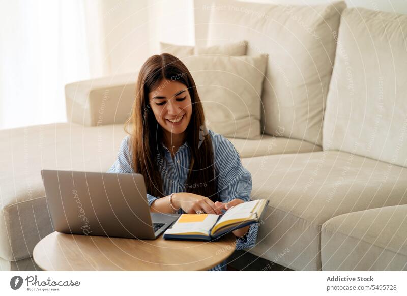 Smiling woman working on laptop in living room entrepreneur freelance read planner smile home remote young female job positive notebook device using online sofa