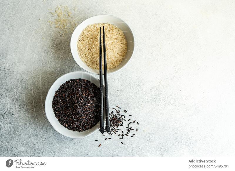 Raw wild black rice and peeled white rice in the bowls with chopsticks. raw ceramic table surface food served menu meal lunch dinner ingredient dish cuisine