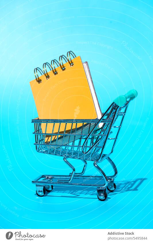 Miniature shopping trolley with hardcovers notebook on blue backdrop shopper cart supermarket miniature spiral concept background store vivid bright creative