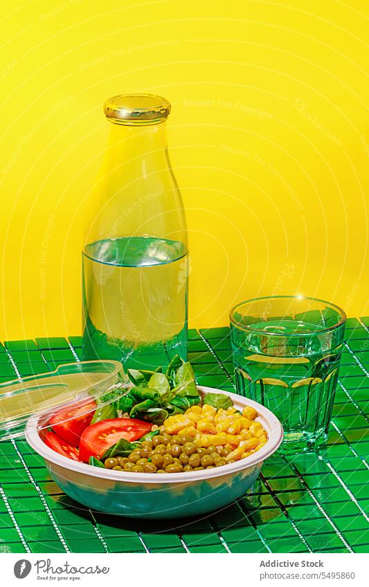 Salad bowl on green background with plastic cover near bottle and glass of water salad food slice tomato spinach leaf corn kernel pea colorful surface table