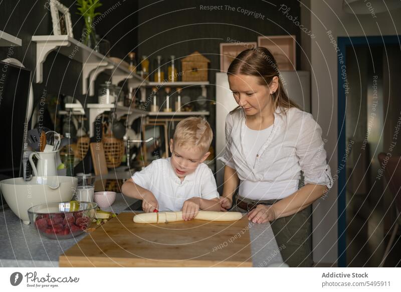 Woman looking at boy cutting banana on board in kitchen mother son cook woman together fruit prepare food family child knife casual attire strawberry cute slice