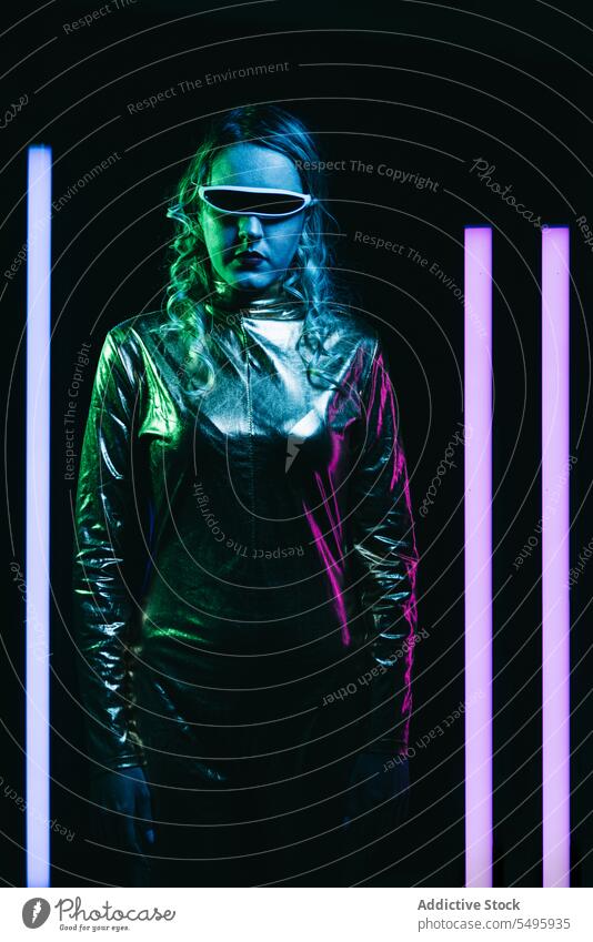 Cybernetic young girl against dark background with lights cybernetic curly hair blonde glasses futuristic wear bright reflecting clothing looking at camera
