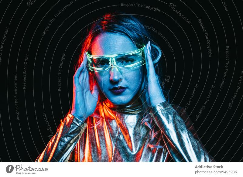 Cybernetic young girl against dark background cybernetic curly hair blonde glasses futuristic wear bright reflecting clothing looking at camera unemotional