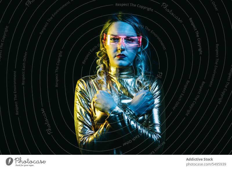 Cybernetic young girl with crossed arms in closed fists against dark background cybernetic curly hair blonde glasses futuristic wear bright reflecting clothing