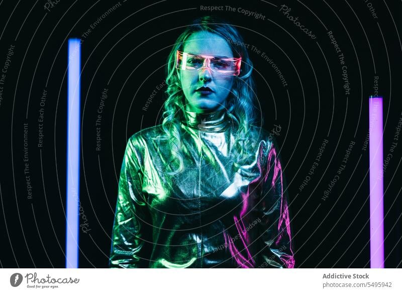 Cybernetic young girl against dark background with lights cybernetic curly hair blonde glasses futuristic wear bright reflecting clothing looking at camera