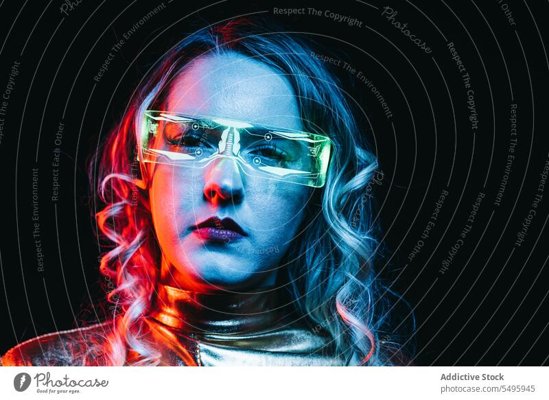 Cybernetic young girl against dark background cybernetic curly hair blonde glasses futuristic wear bright reflecting clothing looking at camera unemotional