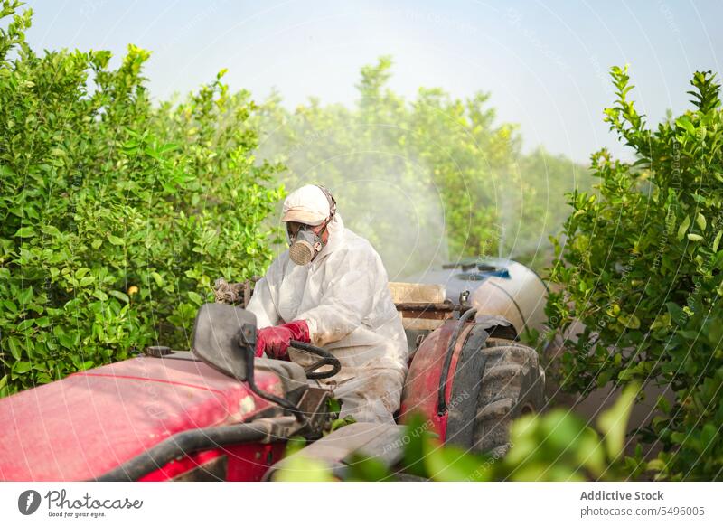 Anonymous farmer spraying pesticide on lemon trees while riding tractor insecticide agriculture protective suit mask chemical nature countryside cultivation