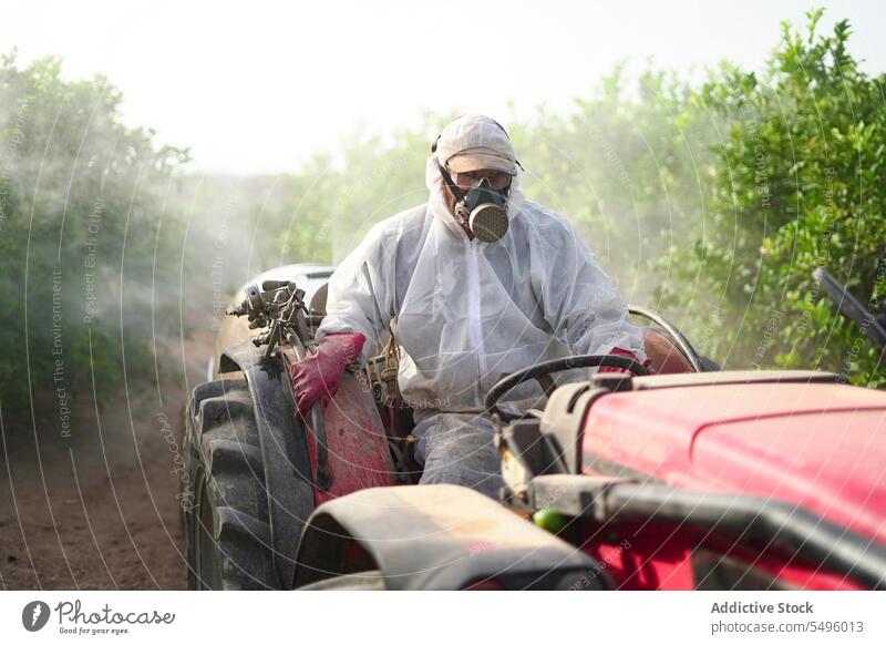 Anonymous farmer spraying pesticide on lemon trees while riding tractor insecticide agriculture protective suit mask chemical nature countryside cultivation