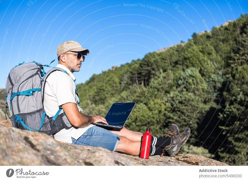 Mature traveler using laptop on rocky cliff man freelance work nature browsing male plant remote gadget summer device internet surfing computer online