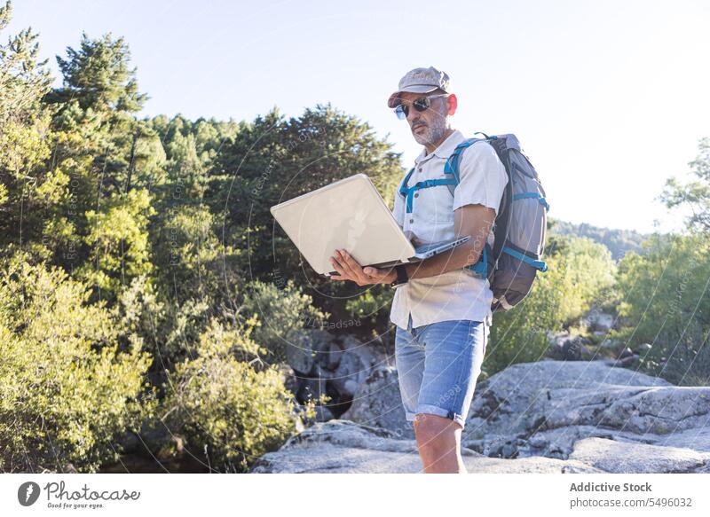 Mature man with backpack using laptop hiker traveler browsing nature rock rocky male journey trip sunglasses gadget adventure summer environment device online