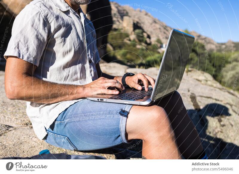 Anonymous crop man using laptop on rocky cliff freelance work nature browsing traveler male plant remote gadget summer device internet surfing computer online